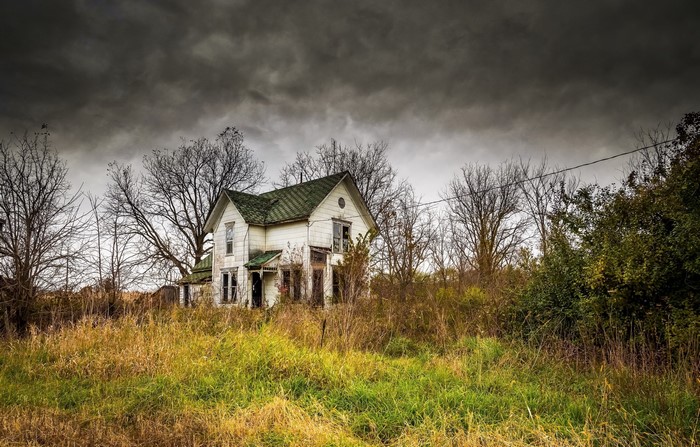 Abandoned house in overgrown field under stormy sky