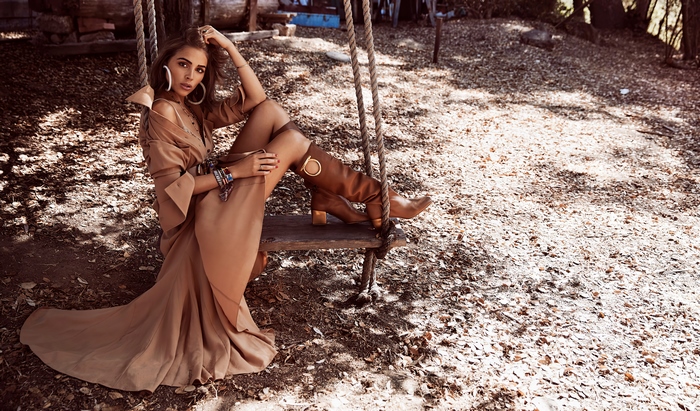 Olivia Culpo posing on a swing in a tan dress and boots