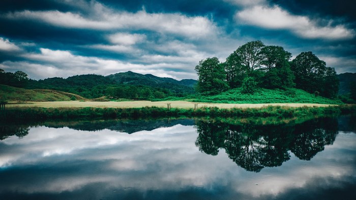 Green mountains and trees reflected in a lake under a cloudy sky