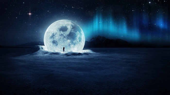 A person stands before a giant moon under the aurora borealis