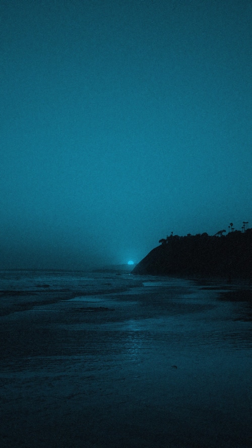Dark ocean waves at dusk with a glowing sun behind a hill