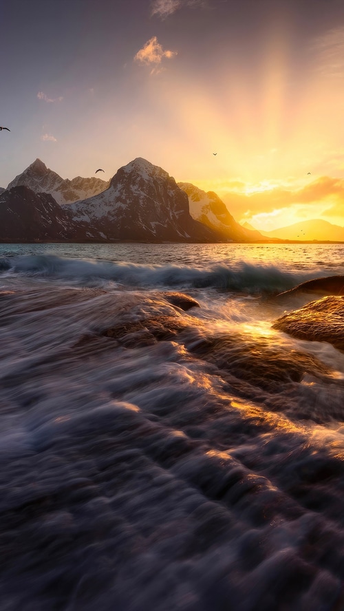 Waves crashing on rocks with snow-capped mountains at sunset