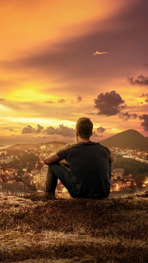 Man sitting on a hill overlooking a city at sunset
