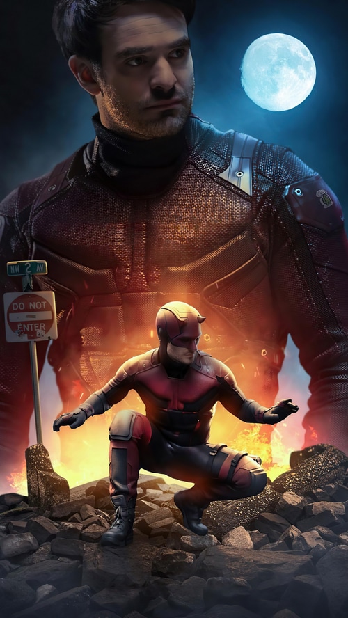 Daredevil in a red suit crouches on rocks with fire behind him