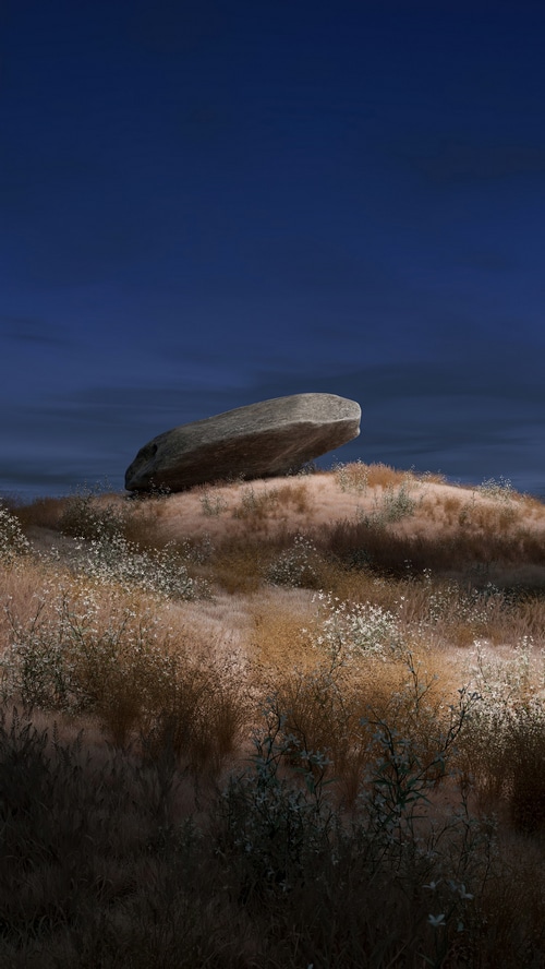 A large rock balanced on a grassy hill under a dark blue sky