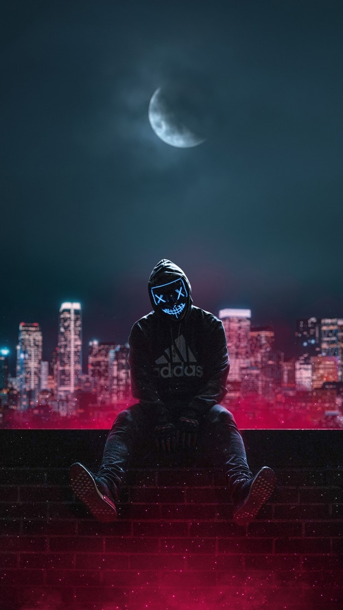 Man in a neon mask sits on a rooftop overlooking a city