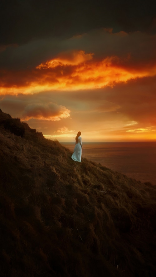 Woman in white dress on grassy cliff overlooking ocean at sunset