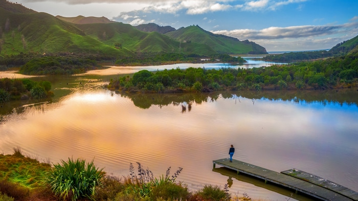 Woman on jetty overlooking water and mountains at sunset
