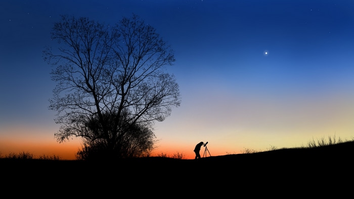 Silhouette of a person stargazing with a telescope under a twilight sky