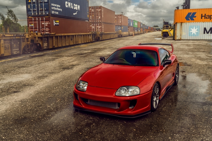 Red Toyota Supra parked near shipping containers and train