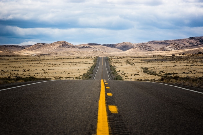A long, empty desert road stretching towards mountains under a cloudy sky.