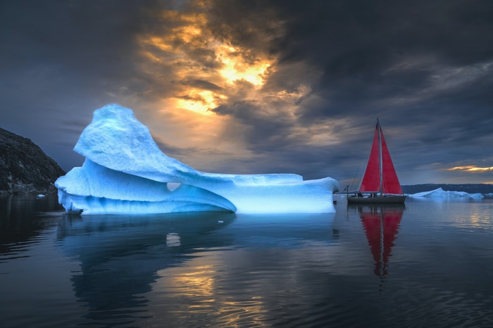 Sailboat with red sail near iceberg at sunset