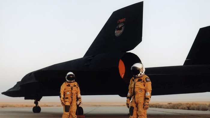 Two pilots in orange suits stand by a Lockheed SR-71 Blackbird