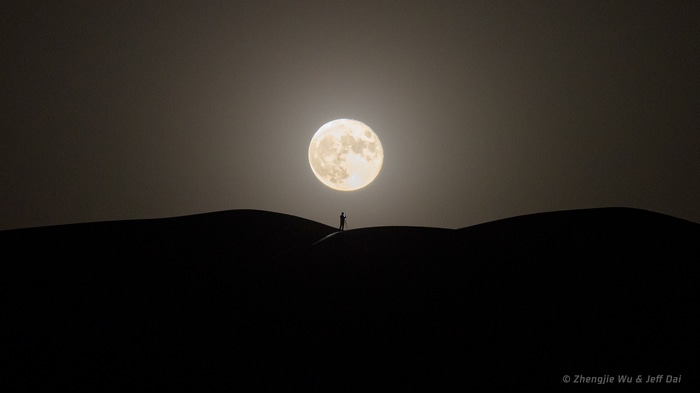Silhouette of a person photographing the Moon over desert dunes