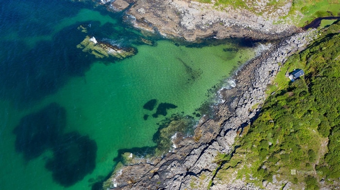 Aerial view of a rocky coastline with clear green water and a small house