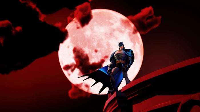 Batman standing on a building ledge with a red moon