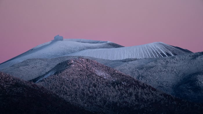 Snow-covered volcano erupting under a pink sky