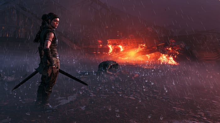 Senua standing in the rain with a burning village