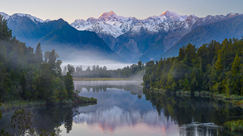 Misty lake reflecting snow-capped mountains and forest