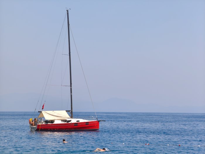 Red sailboat anchored in calm blue sea with swimmers