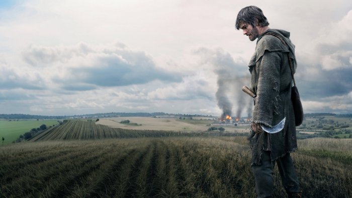 Man with scythe looks at burning village