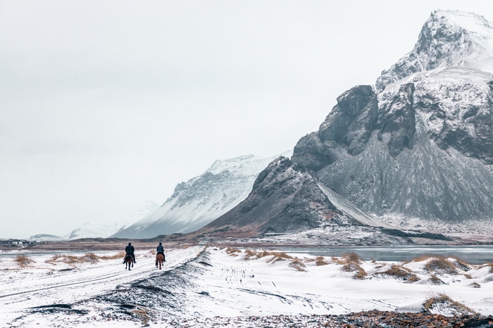 Two people on horseback on a snowy path with mountains