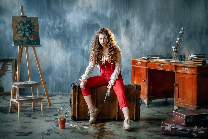 Woman in red overalls sitting on a suitcase with art supplies