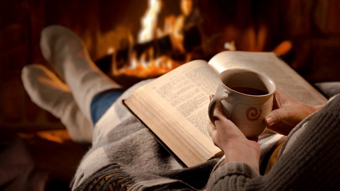 Person reading a book by the fireplace with tea