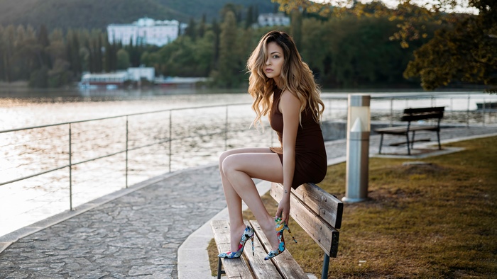 Woman in brown dress and colorful heels sitting on bench by lake
