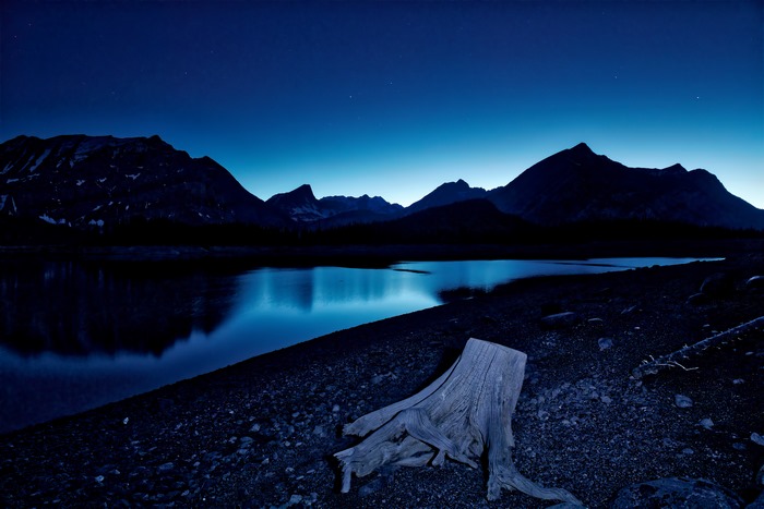 Night sky over mountains and lake with tree stump