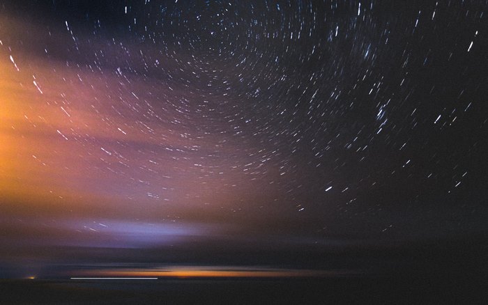 Star trails over a dark landscape with orange and purple clouds