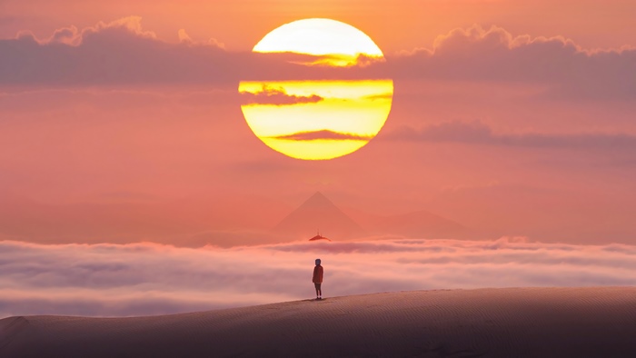Person standing on sand dune at sunset with pyramid and hang glider