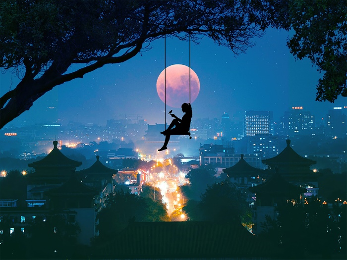 Silhouette of a person on a swing under a large tree with a full moon and city skyline