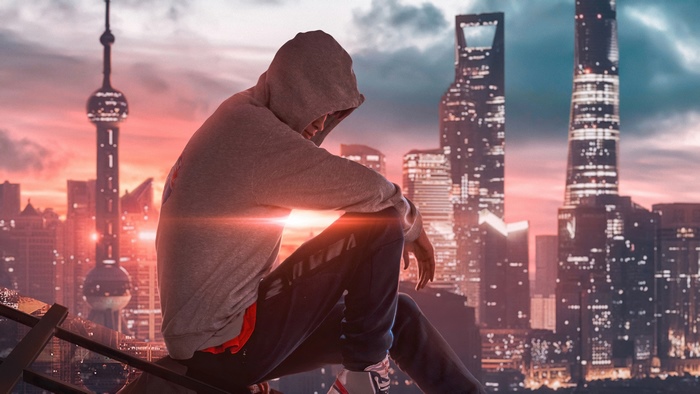 Man in hoodie sitting on a ledge overlooking a city skyline at sunset