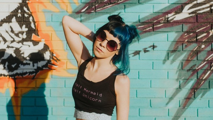 Woman with dyed blue hair and sunglasses in front of graffiti wall