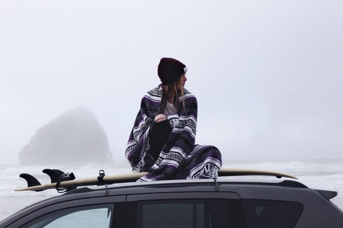 Woman wrapped in blanket on car roof with surfboard