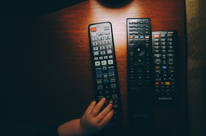 Three remote controls on a wooden table with a child's hand