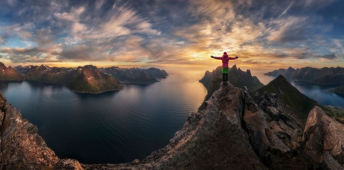 Person on mountain peak overlooking fjord at sunset