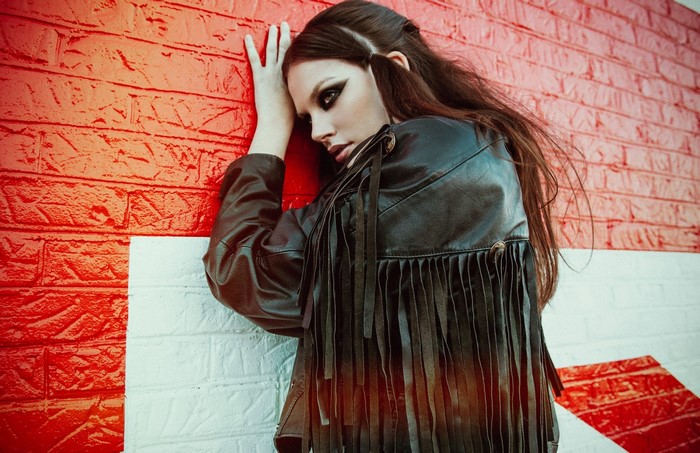 Woman in leather jacket with fringe leaning against brick wall