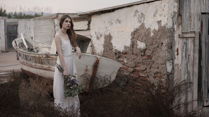 Woman in white dress holding flowers near boat and dilapidated building