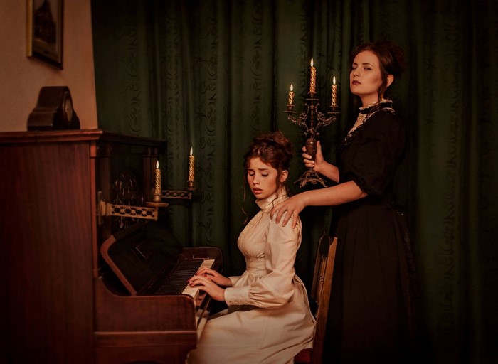 Two women in historical clothing by a piano and candelabra