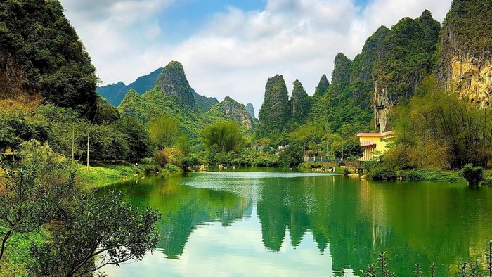 Green lake reflecting karst mountains and trees under a cloudy sky