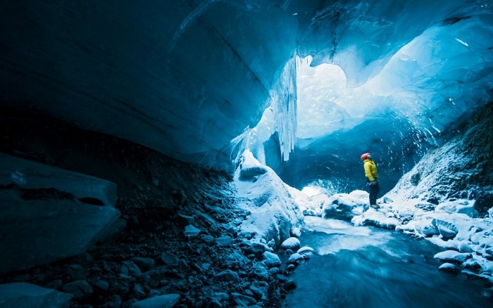 Man in yellow jacket inside a blue ice cave with stream