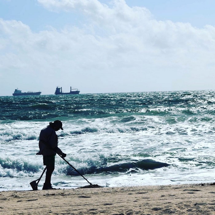 Person metal detecting on a beach with ships in the distance