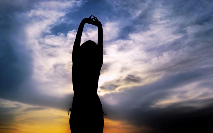 Silhouette of a woman with arms raised against a cloudy sunset sky