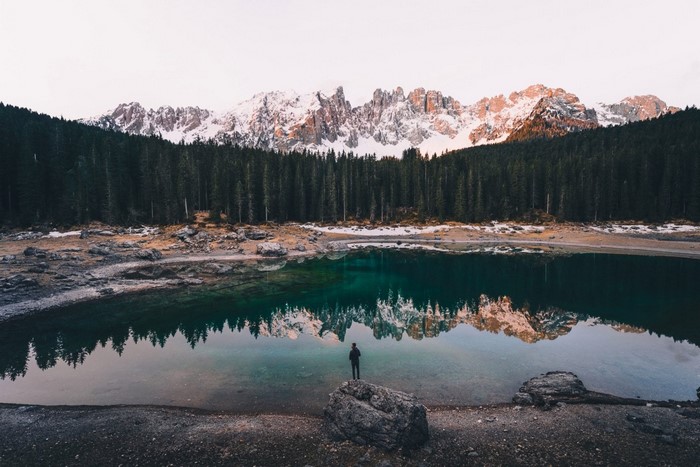 Man standing by a lake with snow-capped mountains and forest reflection