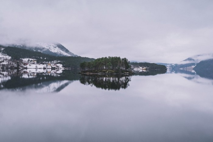 Island with trees reflected in a calm lake with snowy mountains