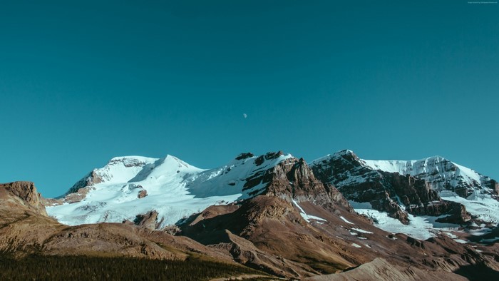 Snow-capped mountains under a clear blue sky with a crescent moon
