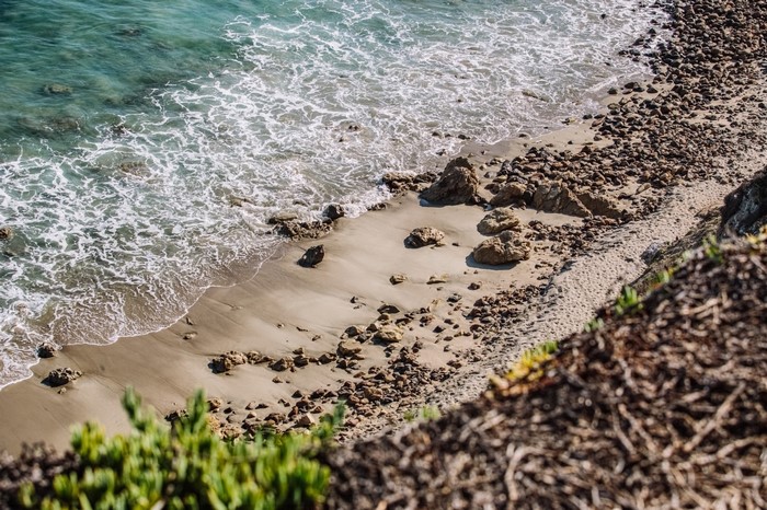 Ocean waves crash on a rocky beach with green foliage in the foreground.
