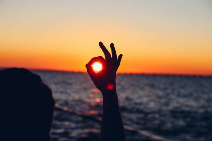 Hand making OK sign with sun at sunset over water